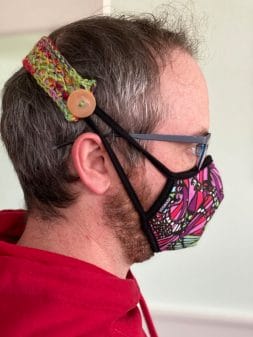 A man's head in profile, demonstrating the use of Ear Savers (a strap with a button), wrapped around his head and connecting to the straps of a protective face mask made of colorful fabric.