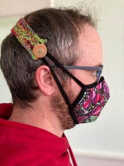 A man's head in profile, demonstrating the use of Ear Savers (a strap with a button), wrapped around his head and connecting to the straps of a protective face mask made of colorful fabric.
