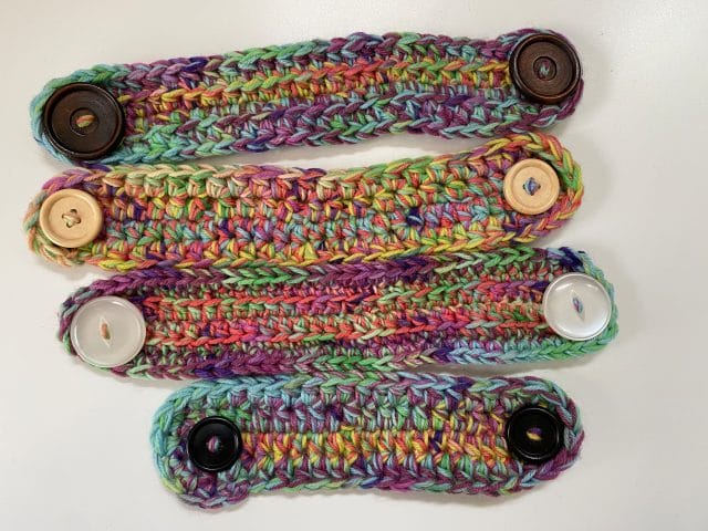 On a white background, a top-down view a pile of crocheted ear savers (4" x 1" rounded rectangles with buttons at each end), made in multicolored yarn.