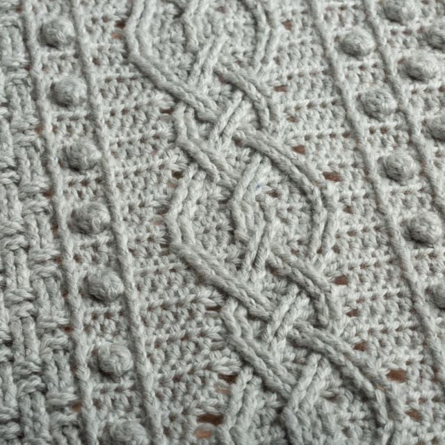 Closeup of the texture of a gray crocheted blanket with intricate cables, bobbles, and basketweave stitch. An impressive one of our free home decor crochet patterns.