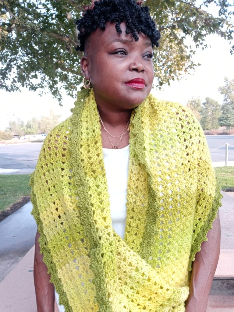 Citrus Wrap by 2 Bossay Knits. A model wears a bright yellow crocheted wrap around her shoulders. It has an avocado green picot edging.