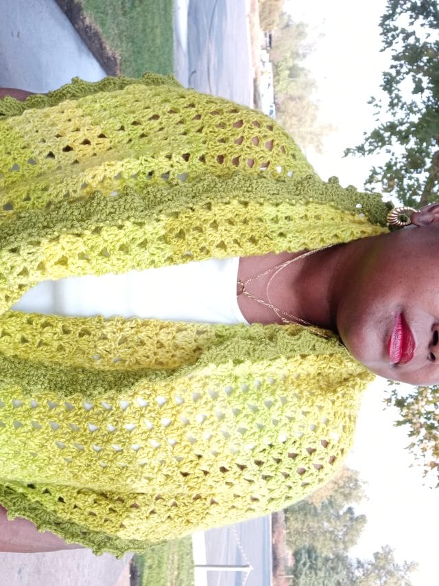 Citrus Wrap by 2 Bossay Knits. A model wears a bright yellow crocheted wrap around her shoulders. It has an avocado green picot edging.