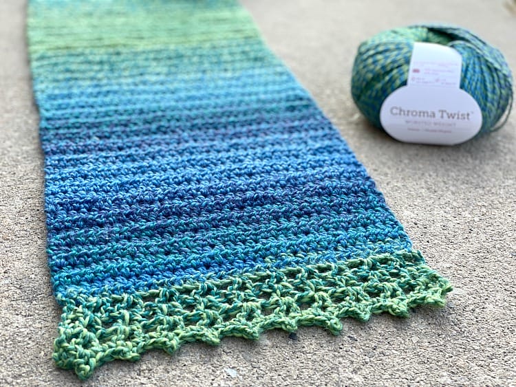 Picot Edged Scarf by 5 Little Monsters. A crocheted blue-green gradient scarf spread out on the ground, with a ball of Chroma Twist in the background.