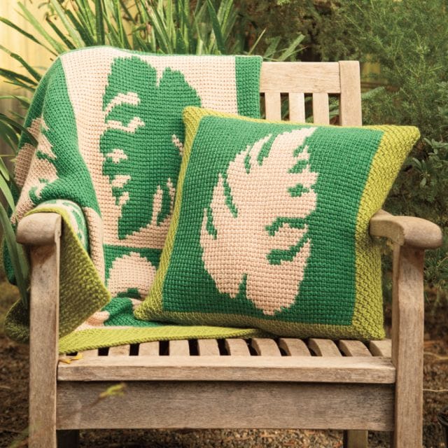 A green and cream blanket and pillow featuring a large monstera leaf motif draped on an outdoor chair. A Tunisian crochet free pattern.