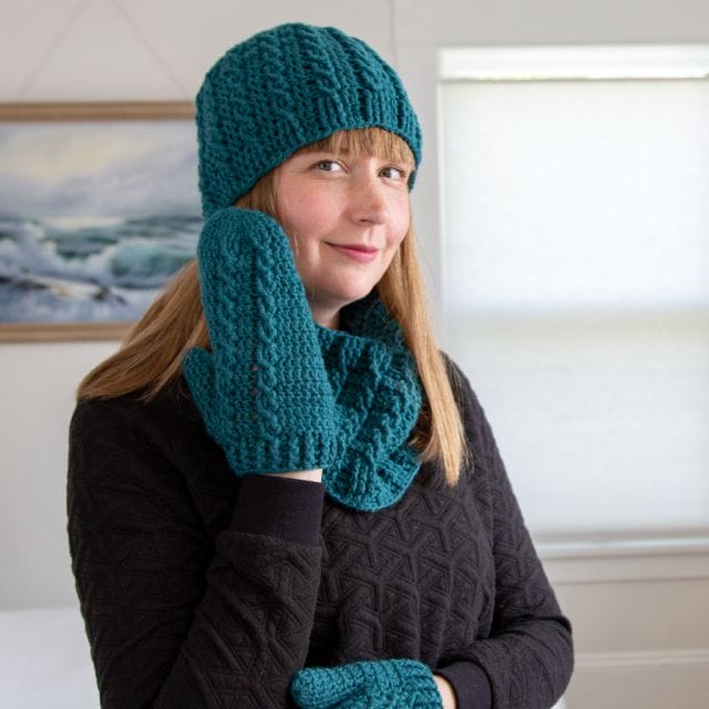 a model wears a crocheted hat, mittens, and cowl