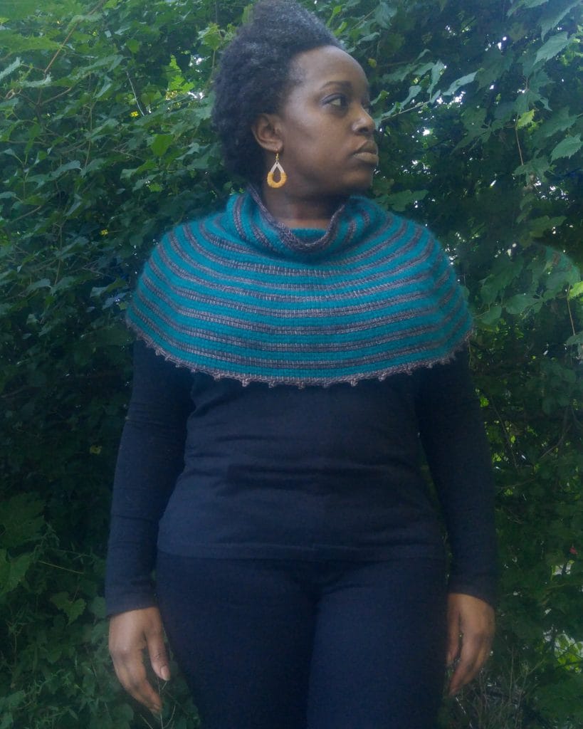 Hayloft Capelet by Detroit Knots. Natalie models the capelet on her shoulders. It is gray and teal striped with a cowl neck.