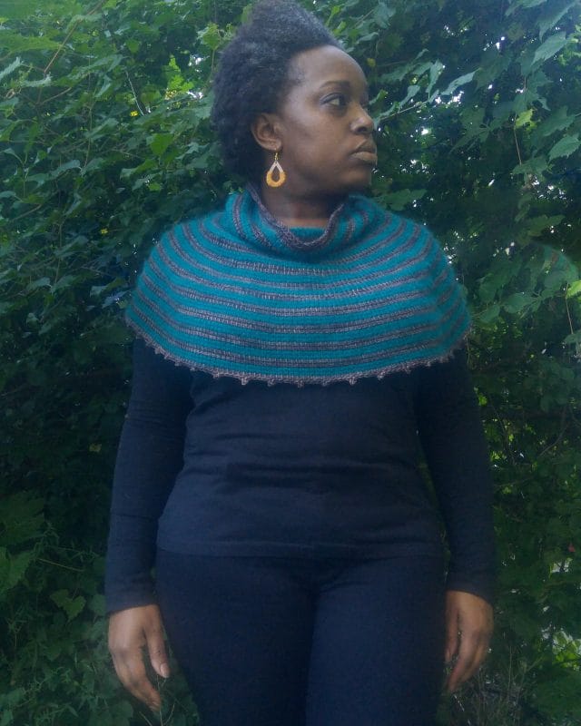 Hayloft Capelet by Detroit Knots. Natalie models the capelet on her shoulders. It is gray and teal striped with a cowl neck.