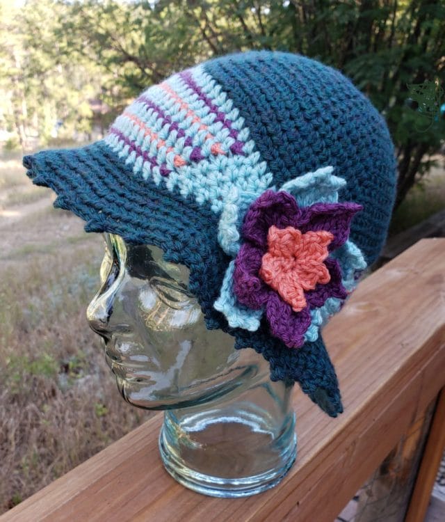 Josephine Cloche by M2H Designs. This crochet hat pattern features a blue hat with a band of blue with purple and pink colorwork and a flower accent.