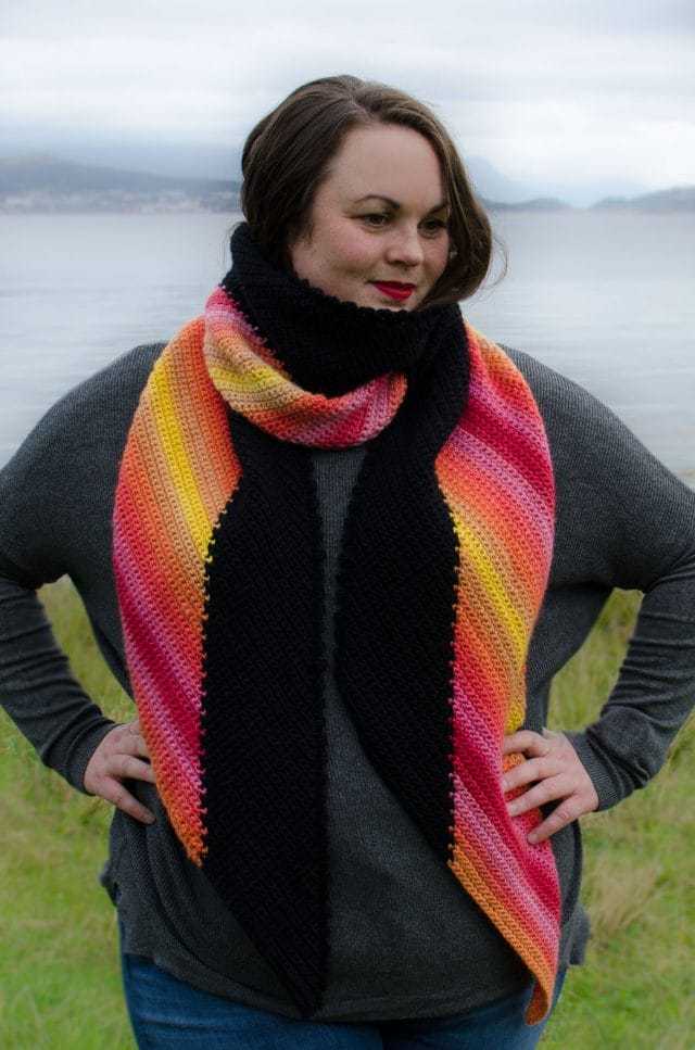 Diagonal Sunset Wrap by Joy of Motion Crochet. Around her neck, a model wears this sunset-colored crocheted scarf edged with black.