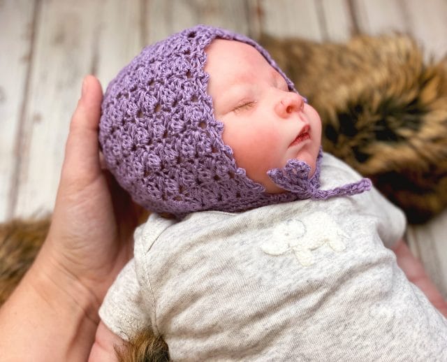 Simple Crochet Baby Bonnet pattern by love.life.yarn. An adult hand touches a newborn baby who is wearing a lavender crocheted baby bonnet.