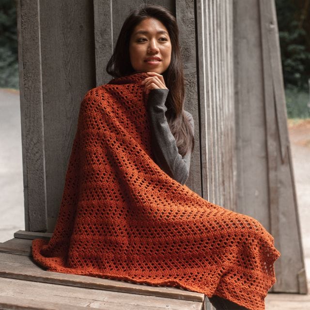 A model is wrapped in the Bambino Ripple Blanket, a copper-colored crochet ripple blanket, part of the free crochet patterns of Crochet.com's 12 Weeks of Gifting.