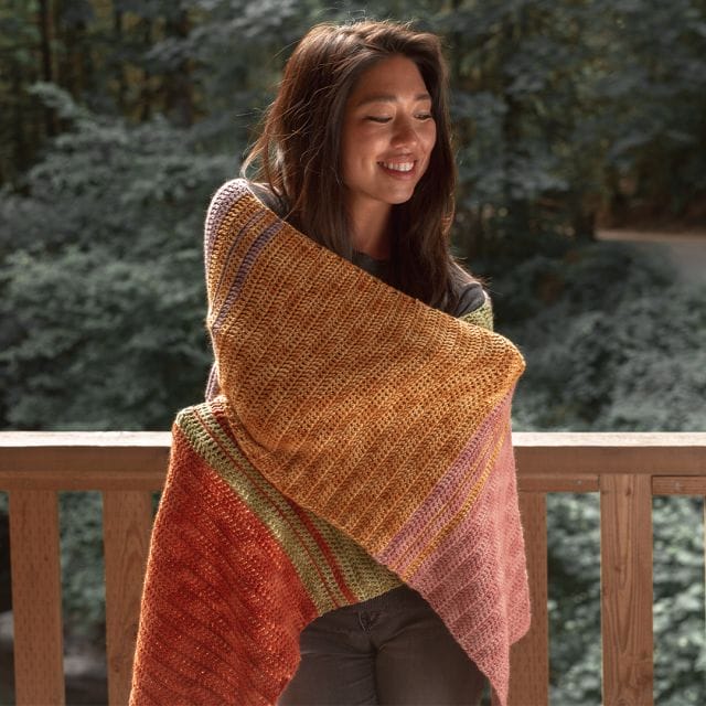 A model is wrapped in the Holiday Sweets Shawl, a multicolored crochet rectangular shawl, part of the free crochet patterns of Crochet.com's 12 Weeks of Gifting.