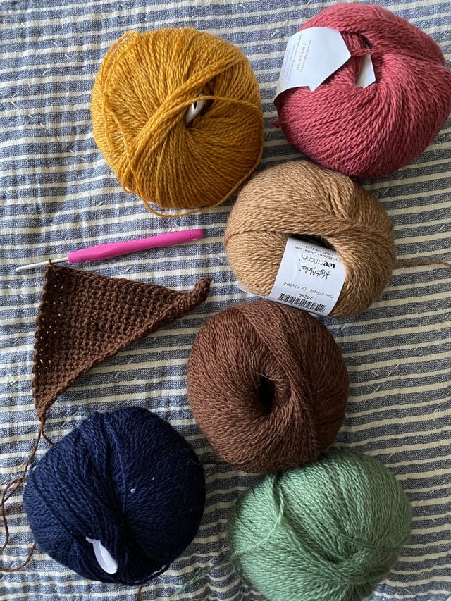 Six balls of Palette yarn in mustard, rouge, tan, brown, sage green, and navy, along with a small triangular crochet swatch and a hook.