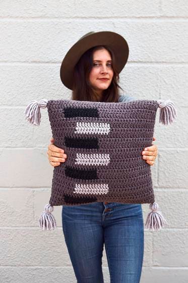 Alexandra holds a finished crocheted pillow, made in gray with white and black rectangle motifs. This is one of our free crochet home decor patterns by Two of Wands.