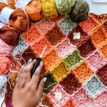 A top-down view of a hand holding a crochet hook against a partially-completed crocheted temperature blanket in orange, rust, and gold tones.