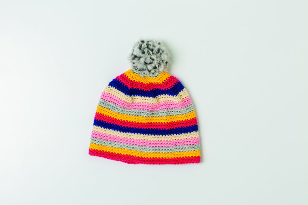 The Brava Beanie, a striped crocheted beanie with a fur pom-pom