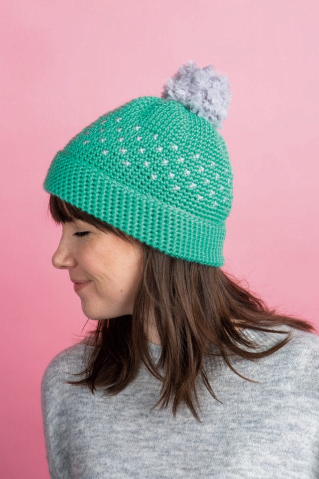A model wears a green crocheted hat with a gray pom-pom