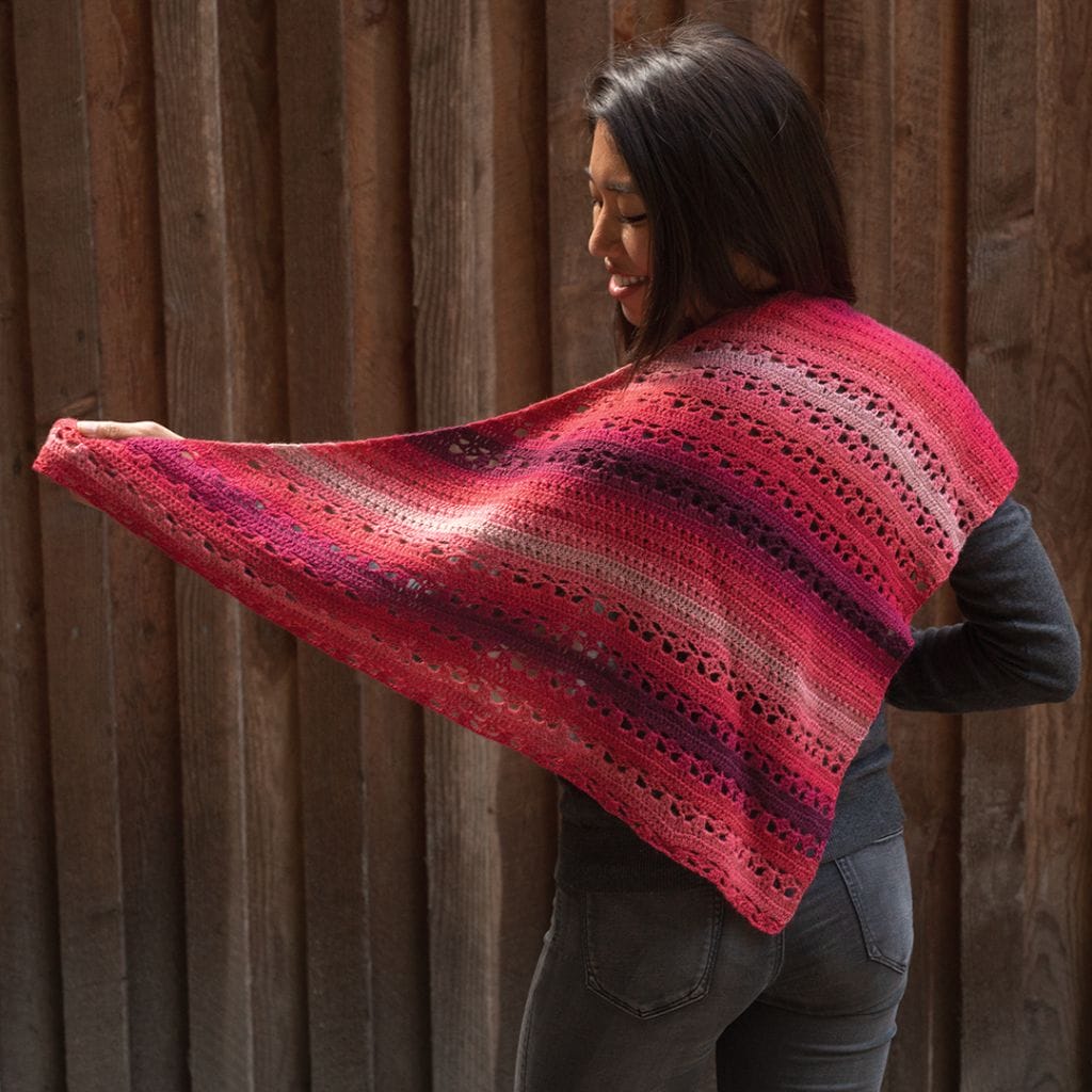 A model wears a red-pink toned crocheted shawl. The Triana shawl is part of the 12 Weeks of Gifting free patterns from Crochet.com