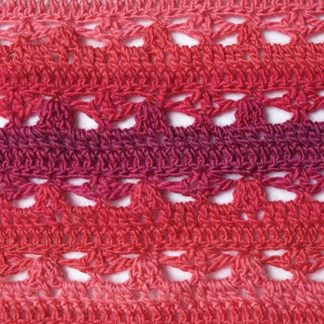 A close up of the stitch pattern in this red-pink toned crocheted shawl. The Triana shawl is part of the 12 Weeks of Gifting free patterns from Crochet.com