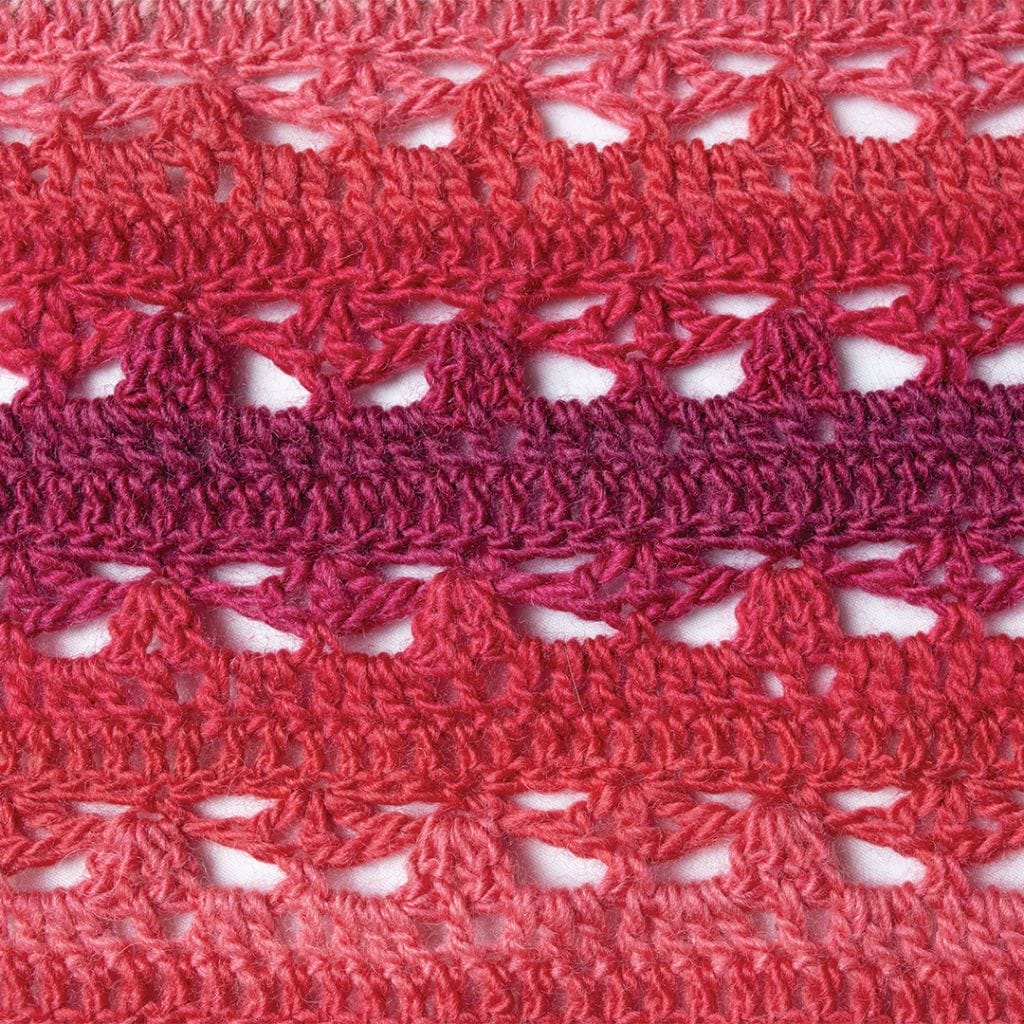 A close up of the stitch pattern in this red-pink toned crocheted shawl. The Triana shawl is part of the 12 Weeks of Gifting free patterns from Crochet.com