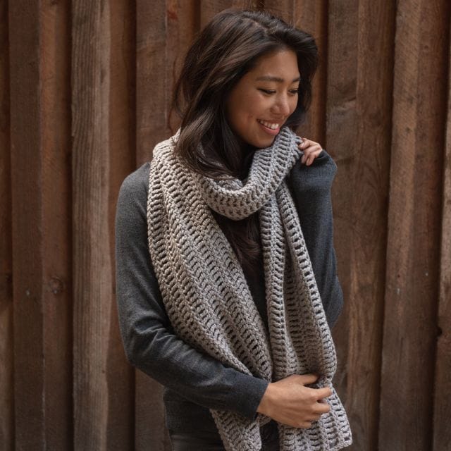A model wears an oversized gray crocheted scarf. The Vanilla Latte Super Scarf, part of the 12 Weeks of Gifting, free crochet patterns from crochet.com