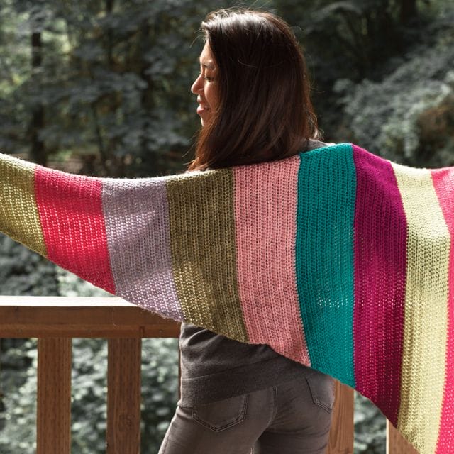 Crochet.com gives away a free crochet pattern every week during the 12 weeks of gifting! This week's pattern is the Bright Gifts Wrap. Image shows: a model wearing a triangular crocheted wrap, shown from behind. The wrap is color blocked in 6" strips of different brightly colored yarn colors, from tan to hot pink, lavender to green, teal to magenta, and bright coral.