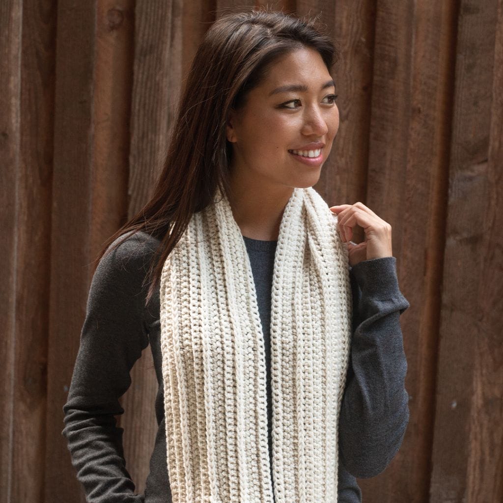 Crochet.com gives away a free crochet pattern every week during the 12 weeks of gifting! This week's pattern is the Cozy Infinity Scarf. Image shows: a model wearing a cream-colored crocheted infinity scarf.