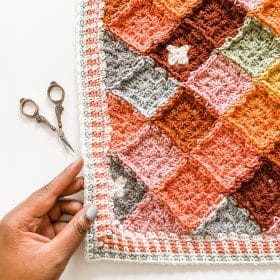 A top-down view of a hand holding the edge of a crocheted temperature blanket in warm colors.