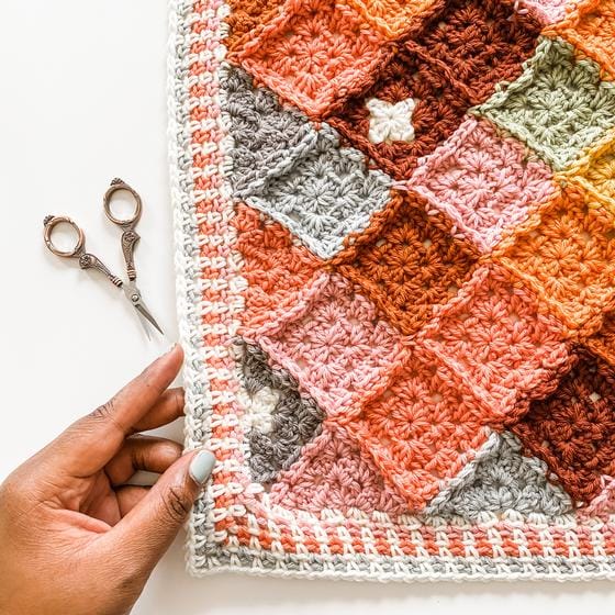 A top down view of a hand holding the corner of a temperature blanket, crocheted in warm tones of orange and coral.