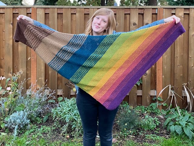 Producer Sarah holds up her finished Faux Fade Shawl, which is a gradient that goes from dark brown to tan to blue to green to yellow to pink to purple.