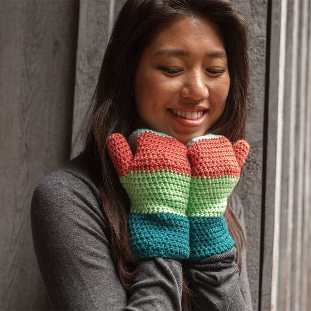 A model wears Mighty Mittens: Crocheted mittens with 4 blocks of color ranging from turquoise, spring green, rust red, and mint green.
