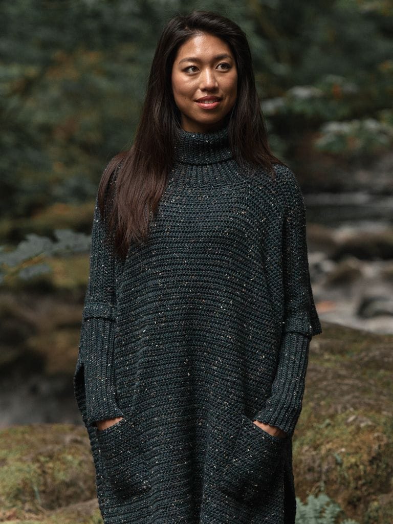 A model wears an oversized gray tweed crocheted poncho. Her hands are in its pockets.