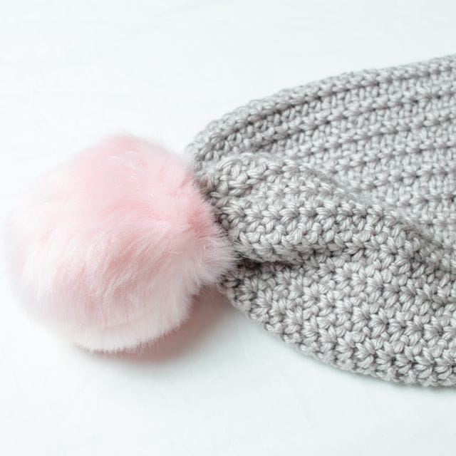 A gray crocheted beanie with a pink pom-pom. The Lisbeth Beanie, a free crochet pattern from crochet.com.