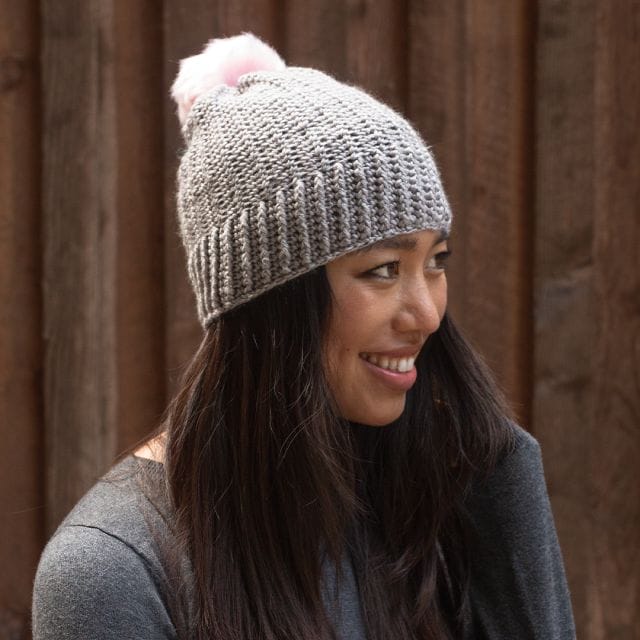 A model wears a gray crocheted beanie with a pink pom-pom. The Lisbeth Beanie, a free crochet pattern from crochet.com.