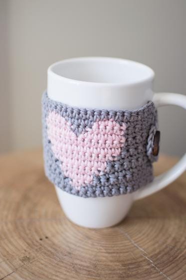 A crocheted mug cozy in gray with a pink heart and fastened with a button