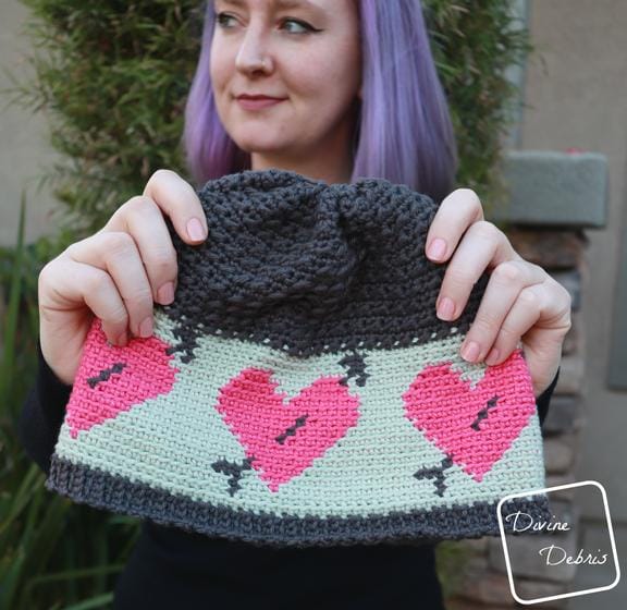 A woman holds a crocheted hat with pink hearts with arrows on it