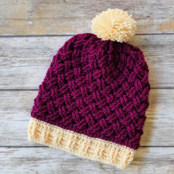 A maroon crocheted hat with criss-cross cables and a cream brim and pom-pom