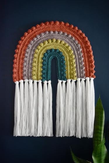 Crocheted rainbow wall hanging in orange, gray, chartreuse, and turquoise, with long white fringe. Check crochet.com for the best crochet patterns!