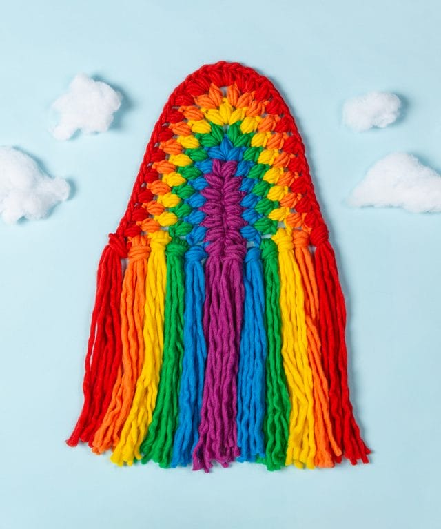 A crocheted rainbow wall hanging on a blue background with clouds made out of stuffing. One of the free crochet patterns from crochet.com!
