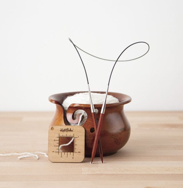 A dark wood yarn bowl with a white ball of yarn inside, along with a gauge swatch tool and a set of knitting needles
