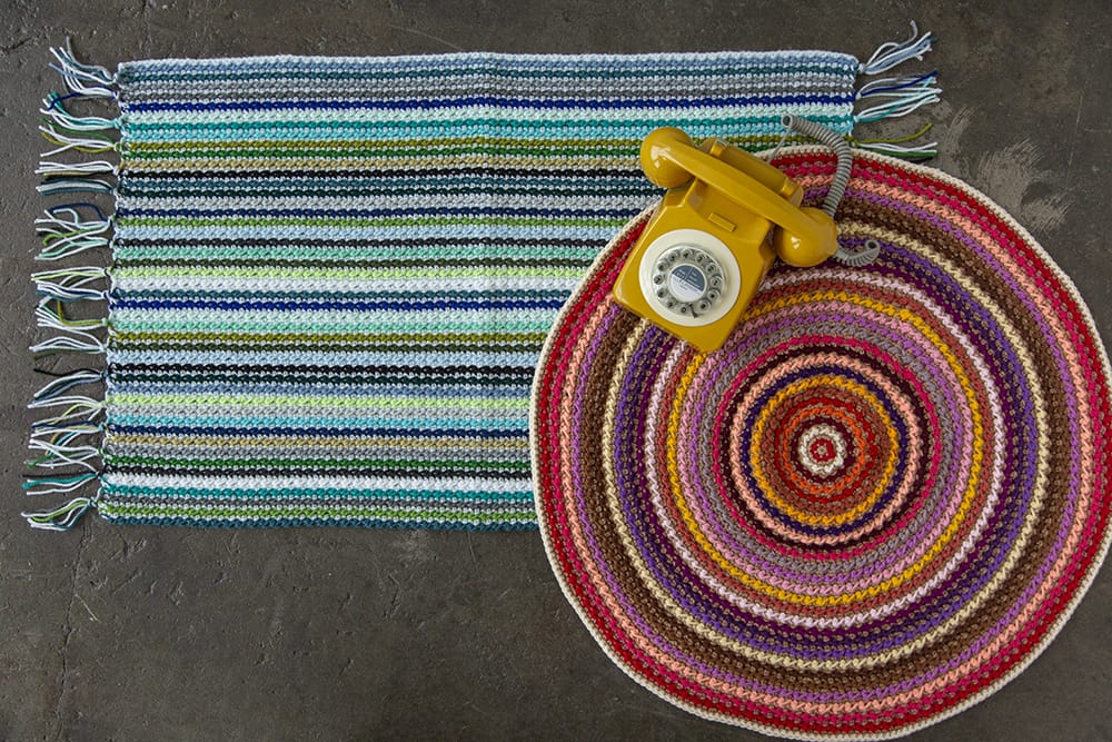 Clean Sweep Rug crochet pattern: A top-down view of a rectangular multicolored striped rug in blue and green tones, with fringe on the ends. Layered on top of it to the right is a round multicolored rug in red/purple tones. A yellow rotary phone is sitting on top of the round rug.