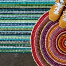 Clean Sweep Rug crochet pattern: A top-down view of a rectangular multicolored striped rug in blue and green tones, with fringe on the ends. Layered on top of it to the right is a round multicolored rug in red/purple tones. A pair of yellow retro running shoes is sitting on top of the round rug.