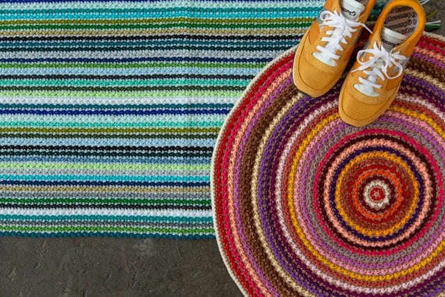 Clean Sweep Rug crochet pattern: A top-down view of a rectangular multicolored striped rug in blue and green tones, with fringe on the ends. Layered on top of it to the right is a round multicolored rug in red/purple tones. A pair of yellow retro running shoes is sitting on top of the round rug.