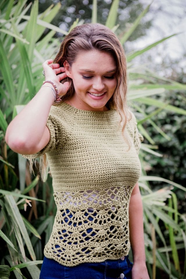 Bahama Blouse by Crochet Foundry. A woman wears a crocheted t-shirt made from straw-colored yarn. It has a solid top and a lacy bottom, beginning just under the bust.