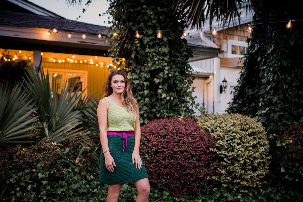 Baybreeze Dress by Crochet Foundry: In a garden, a woman wears a crocheted tank dress with a light green tank top-style top, a shocking purple waistband with drawstring, and a dark green skirt.