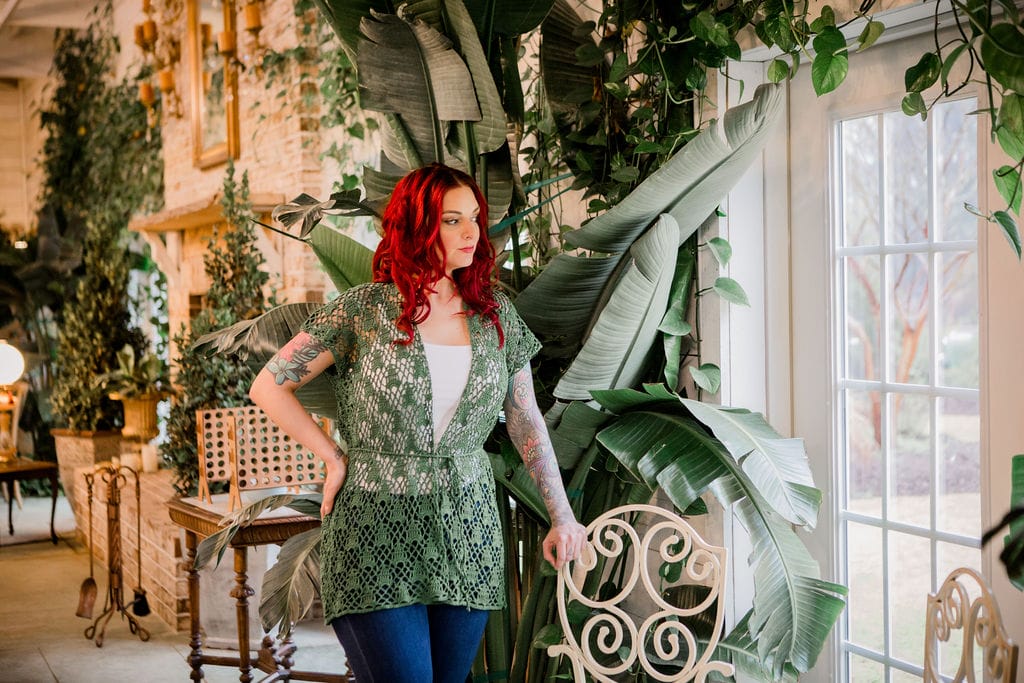 Bora Bora Cardigan by Michelle Moore: In a room full of plants, a woman wears a lacy green crocheted cardigan with triangular motifs and a narrow string belt tied at the waist.