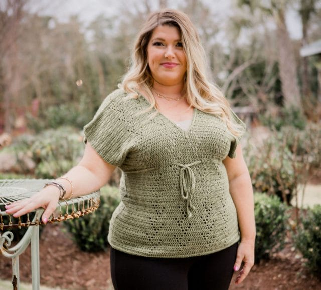 Verona tee by Crochet Foundry. A woman wears a light green crocheted V-neck top with a draw string under the bust, and an openwork pattern repeating down the front.