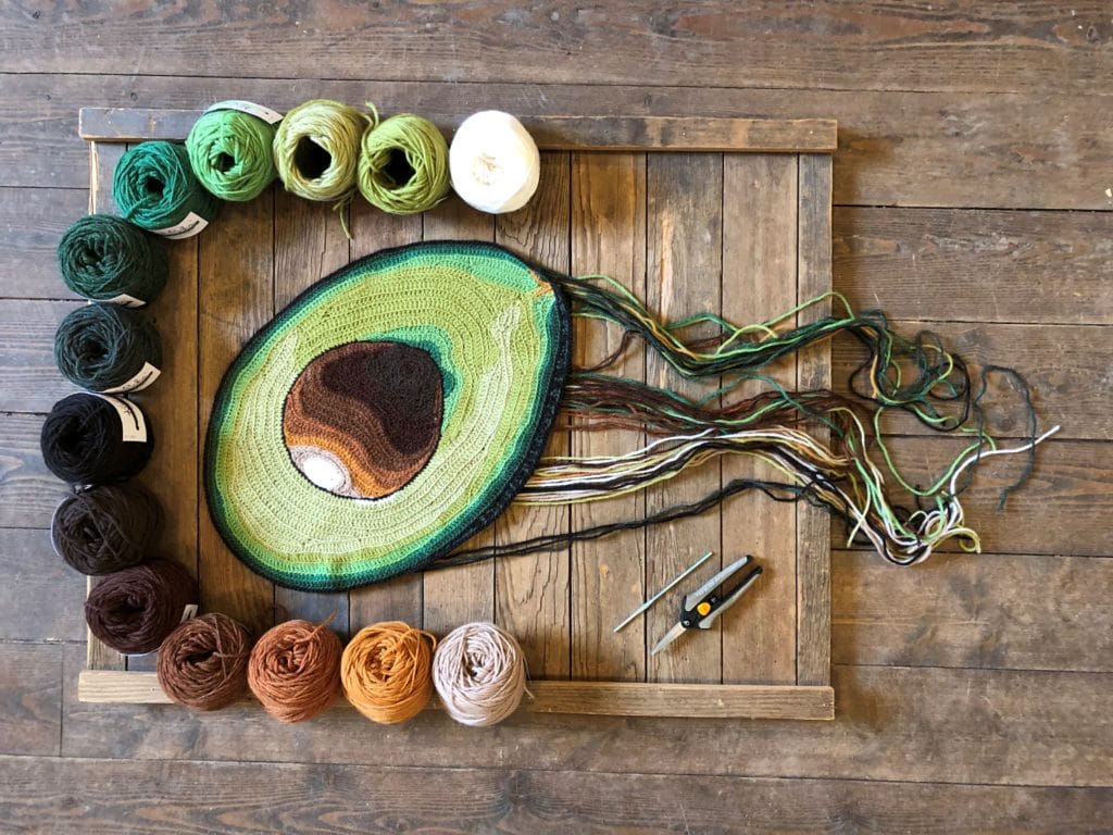 Pieces from the Guacamole series, a 2-D series of hand-crocheted pieces featuring components of guacamole, each piece has a trail of ends emanating from it and is surrounded by a row of yarns in its color palette. 1. A hand-crocheted half an avocado, surrounded by the yarn color palette ranging from greens to brown to golds.
