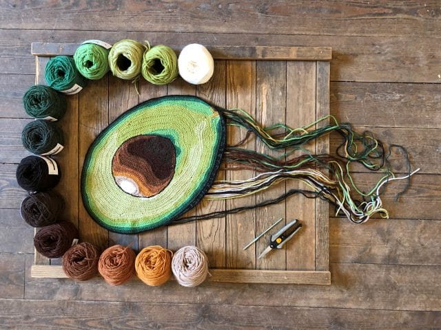 Pieces from the Guacamole series, a 2-D series of hand-crocheted pieces featuring components of guacamole, each piece has a trail of ends emanating from it and is surrounded by a row of yarns in its color palette. 1. A hand-crocheted half an avocado, surrounded by the yarn color palette ranging from greens to brown to golds.