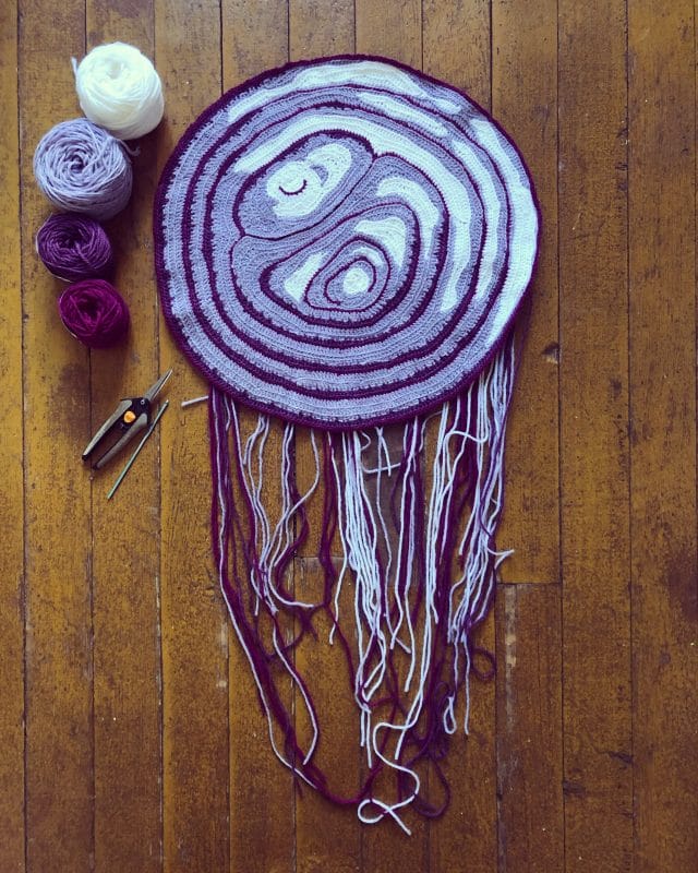 A hand-crocheted art piece by Kate Moran: slice of red onion surrounded by yarn in white and a range of purples.